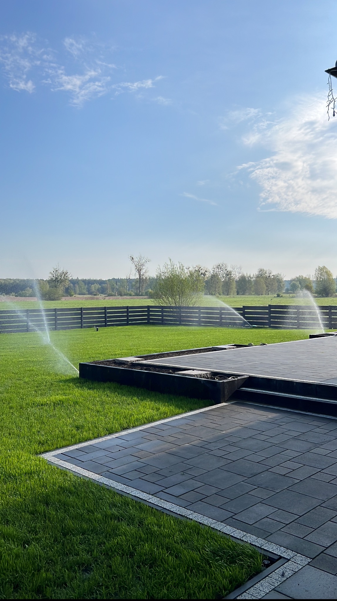 Zraszanie trawnika z systemem nawadniania, widok na równo przystrzyżoną trawę, szarą kostkę brukową i drewniane ogrodzenie na tle błękitnego nieba.