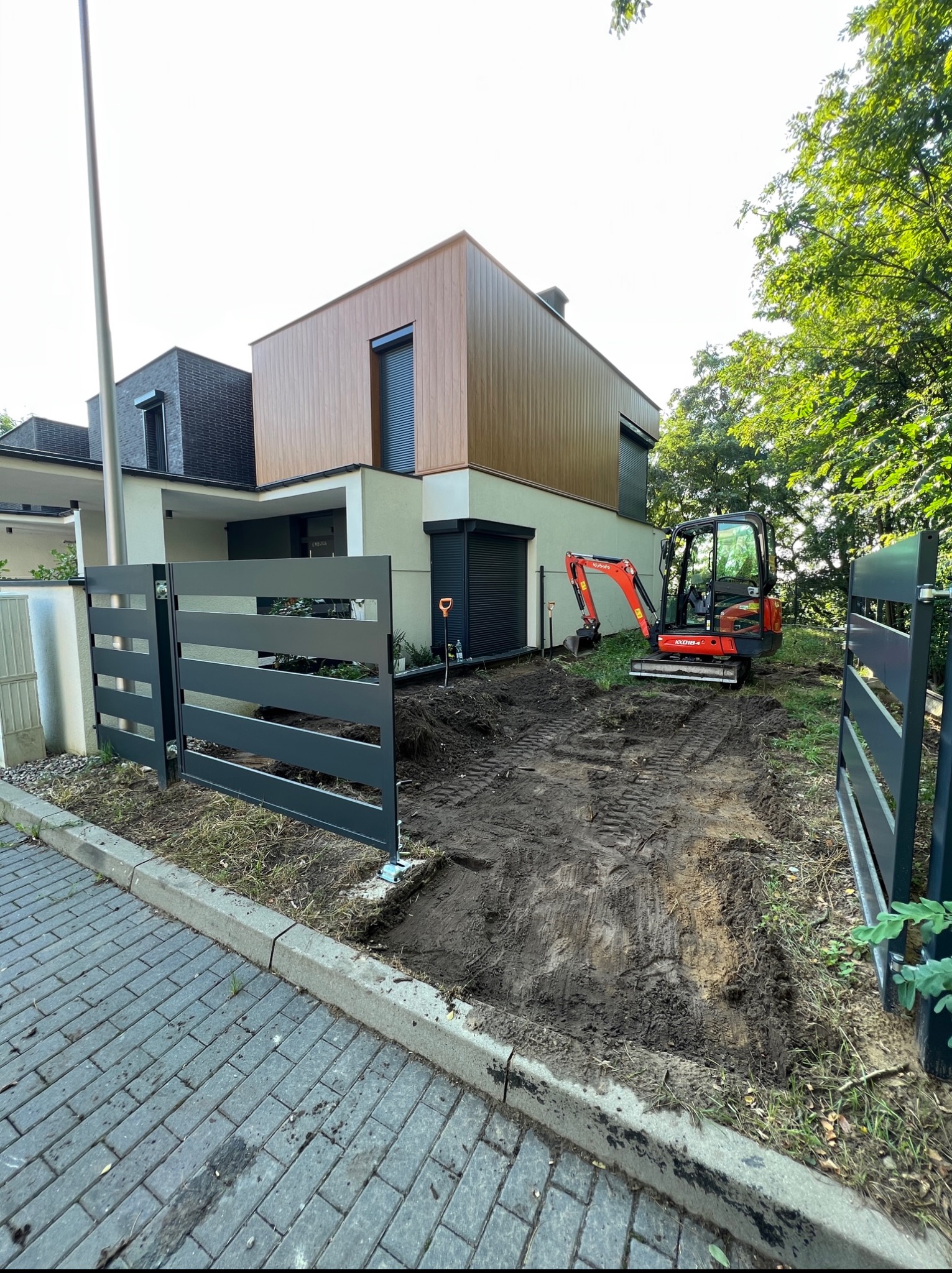 Niwelacja terenu podjazd do nowoczesnego domu. Widoczny minikoparka, ogrodzenie panelowe i wykop pod przyszły podjazd. Słoneczny dzień.