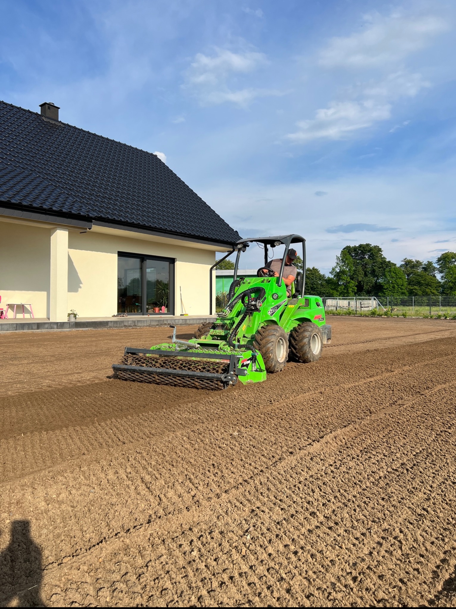Zielony traktor z wałem ugniata ziemię na wyrównanym terenie przed domem. Przygotowanie pod trawnik w słoneczny dzień.