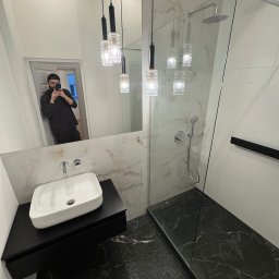 Oferteo A.H - Nowoczesna łazienka z czarnym marmurem na podłodze i ścianach, kabina prysznicowa ze szklaną ścianą, biała umywalka nablatowa i lampy wiszące. Odbicie osoby w lustrze.