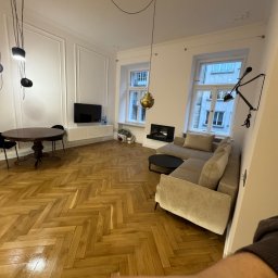 Oferteo A.H - Eleganckie wnętrze po remoncie: parkiet w jodełkę, beżowa sofa, okrągły stół i designerskie lampy. Widok z perspektywy osoby trzymającej telefon.