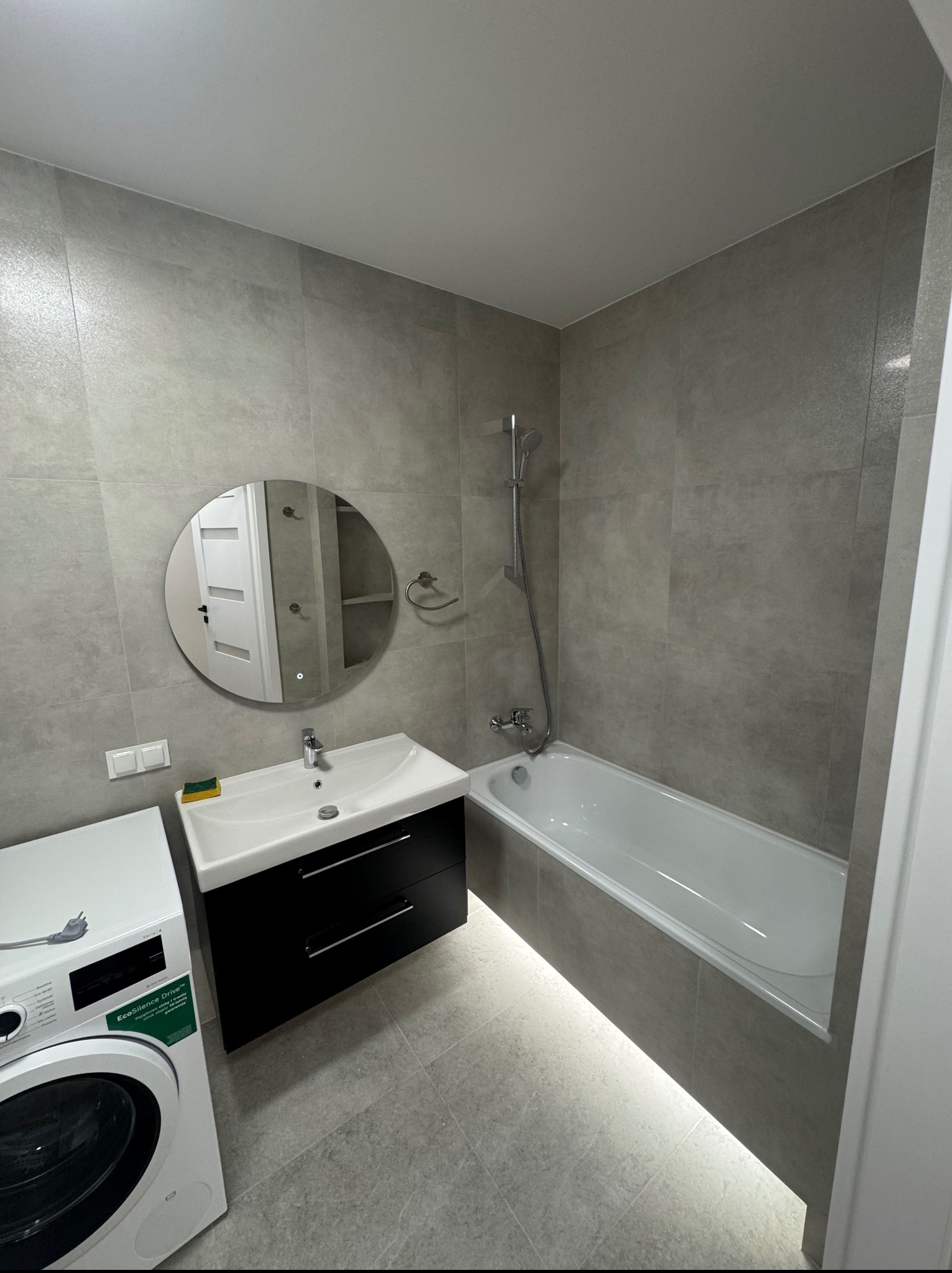 Nowoczesna łazienka w szarościach z podświetloną wanną, okrągłym lustrem i pralką. Minimalistyczny design i stonowana kolorystyka.