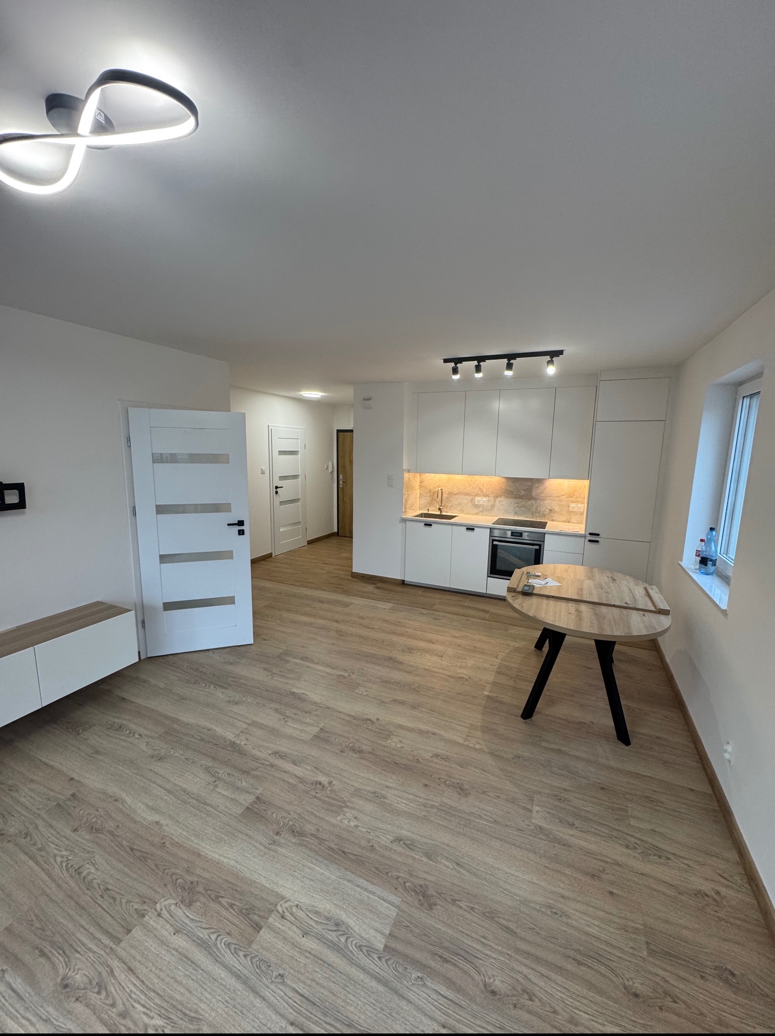 Jasne wnętrze mieszkania po remoncie: panele podłogowe, białe ściany, nowoczesne oświetlenie, zabudowa kuchenna z oknem. Widok na korytarz i inne pomieszczenia.