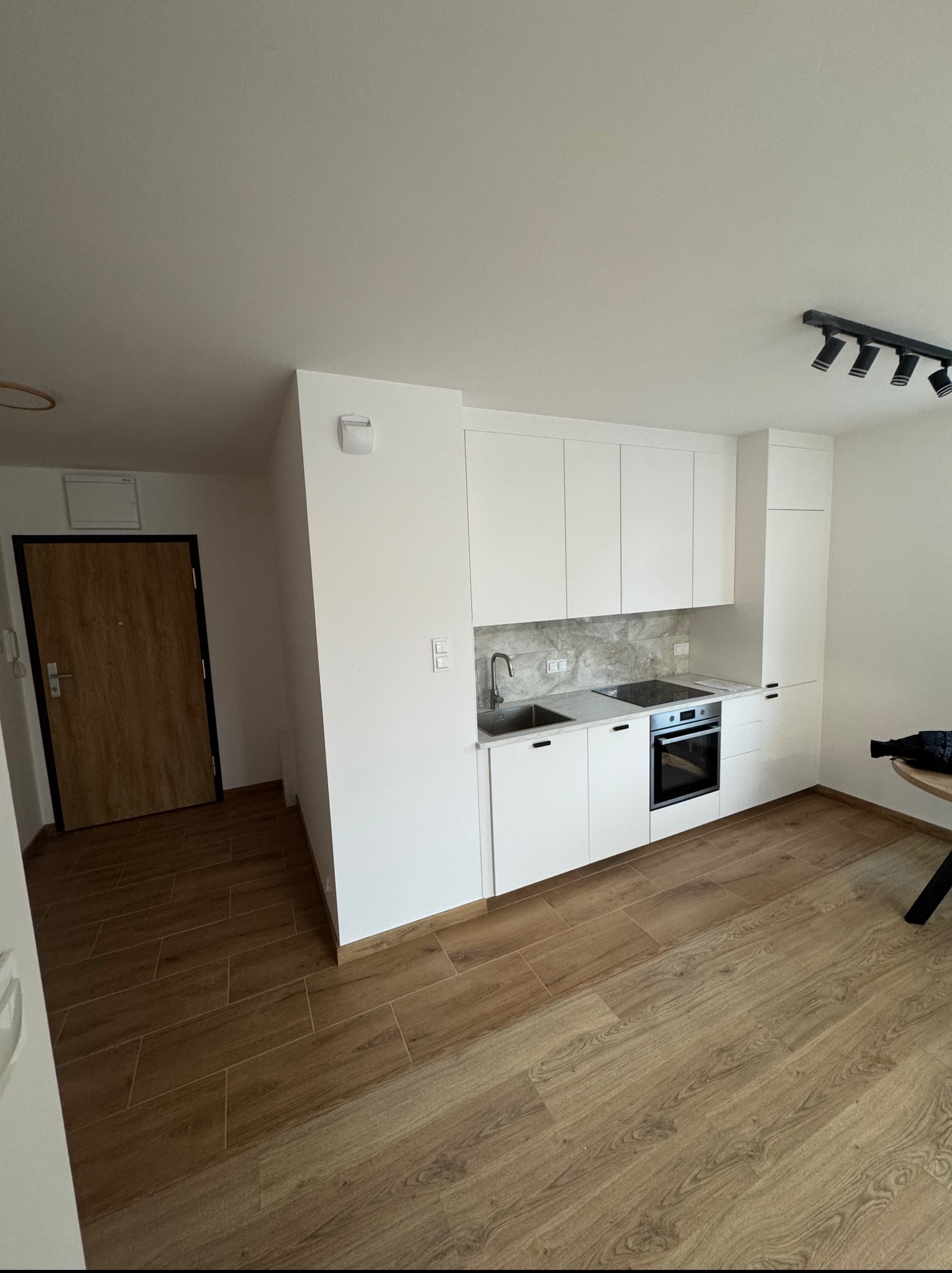 Jasna kuchnia w nowoczesnym stylu z białymi szafkami, drewnianą podłogą i czarnymi reflektorami sufitowymi. Minimalistyczny design, świeżo po remoncie.