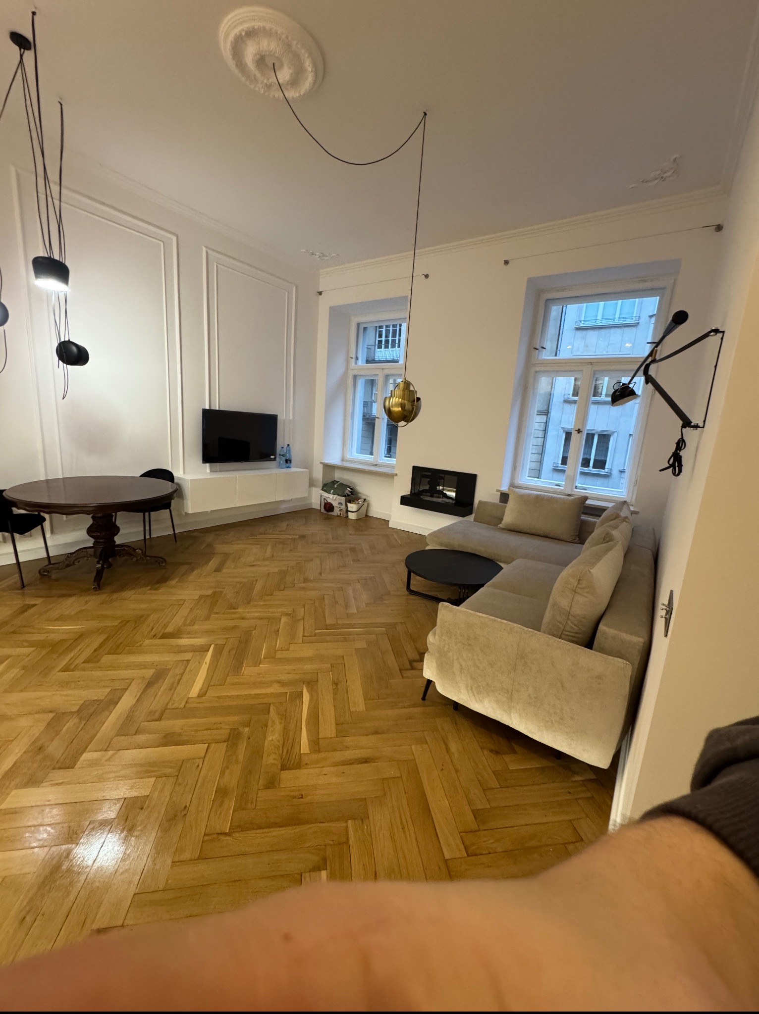 Eleganckie wnętrze po remoncie: parkiet w jodełkę, beżowa sofa, okrągły stół i designerskie lampy. Widok z perspektywy osoby trzymającej telefon.