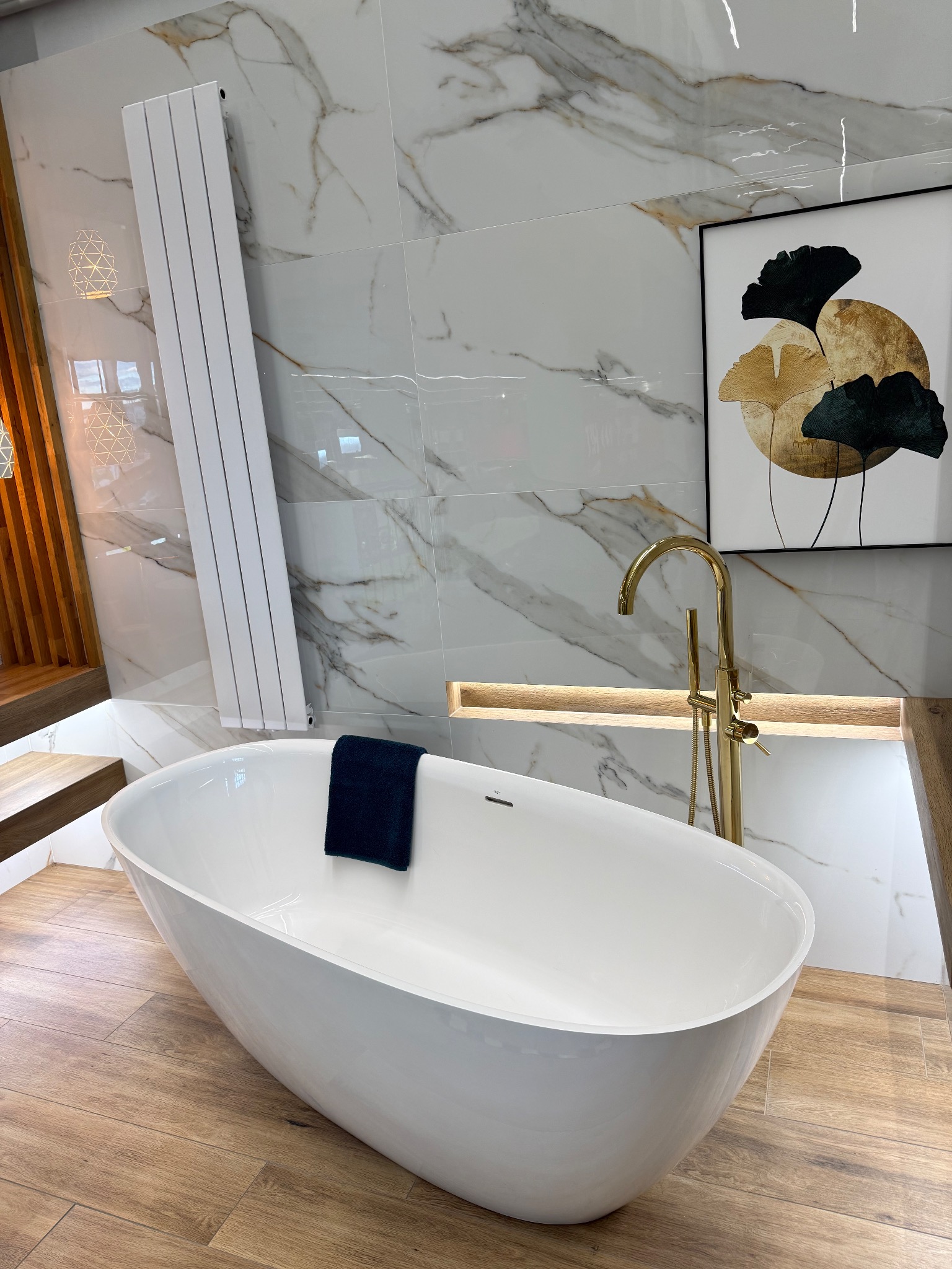 Elegancka, wolnostojąca wanna owalna z baterią w kolorze złota, ręcznikiem i grzejnikiem na tle marmurowej ściany i drewnopodobnej podłogi. Całość dopełnia obraz w czarnej ramie.