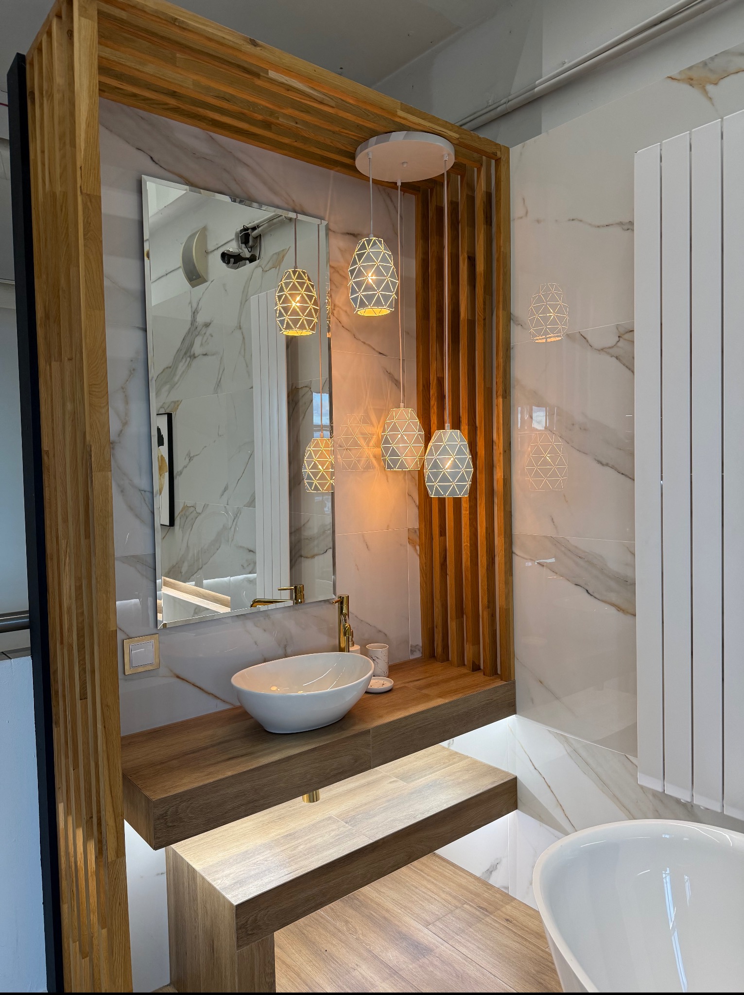 Nowoczesna łazienka z marmurowymi płytkami, drewnianą zabudową umywalki i dekoracyjnym oświetleniem. Lustro odbija wnętrze łazienki, a wiszące lampy dodają elegancji.