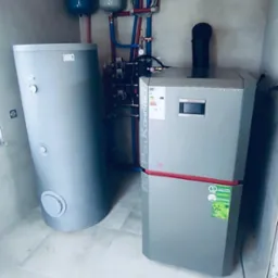 Zestaw kotła na pellet i zasobnika w kotłowni w Łodzi. Widoczne rury instalacji CO i CWU. Kompaktowy system grzewczy w jasnym pomieszczeniu.