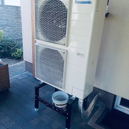 UniEnergy Emilia Szczawińska - Biała pompa ciepła Panasonic na czarnym stojaku, obok ściany budynku z oknem. Widoczny fragment kostki brukowej i roślinności w tle.