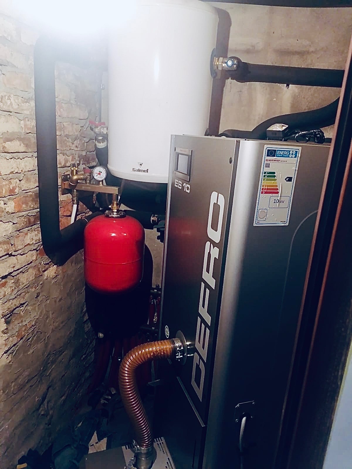 Nowoczesny piec Defro ES 10 z zasobnikiem Galmet w kotłowni z ceglaną ścianą. Widoczny manometr, przewody i czerwony zbiornik wyrównawczy.