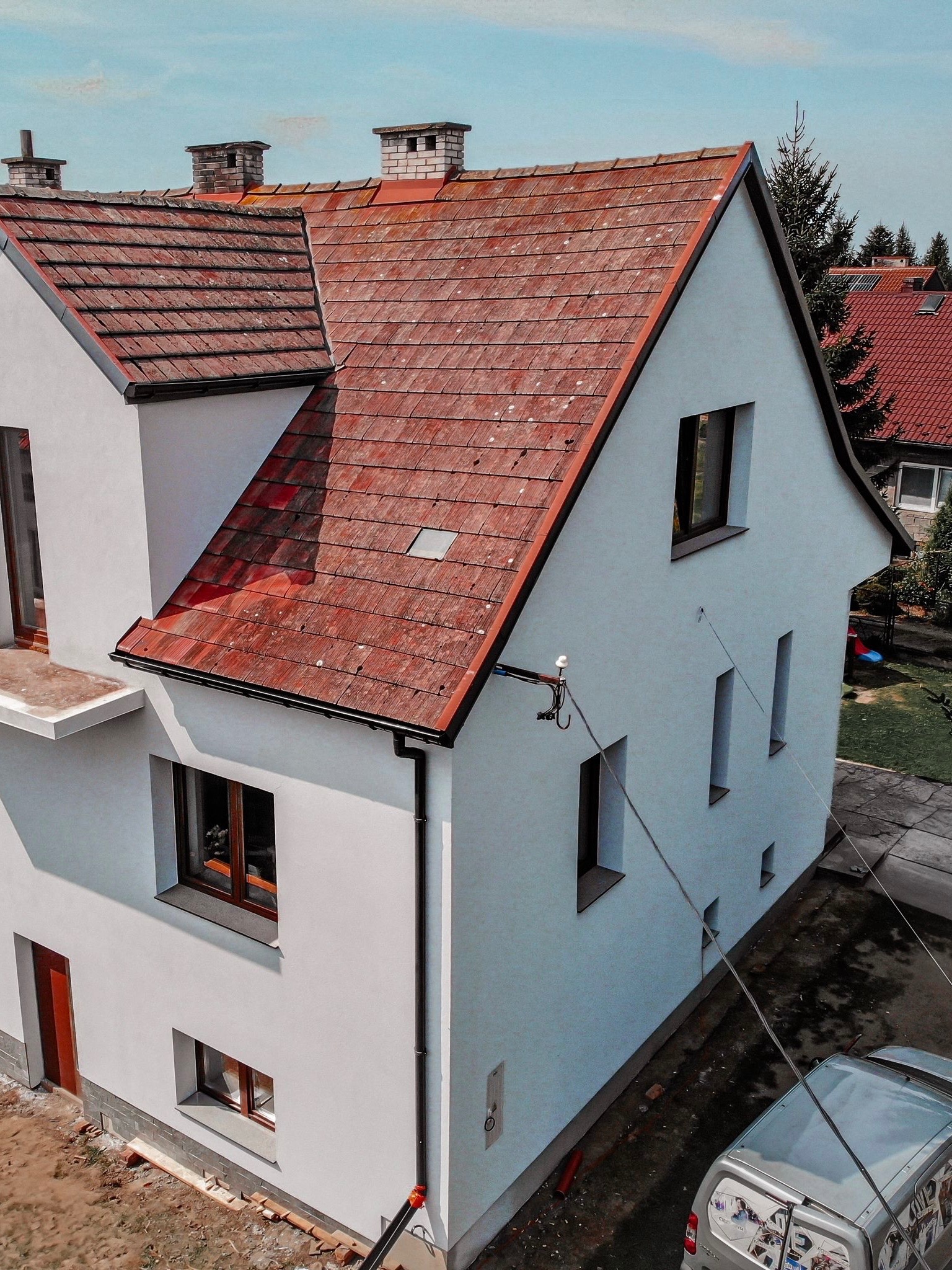 Biały dom jednorodzinny z czerwoną dachówką i trzema kominami. Widoczne okna z brązowymi ramami, rynna i fragment instalacji na elewacji. Ujęcie z góry, częściowo widoczny samochód.