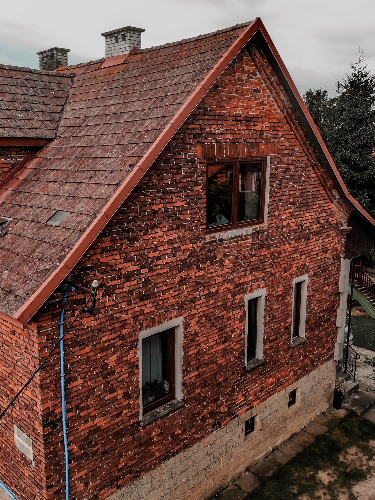Ceglany dom jednorodzinny z czerwonym dachem, widoczny z góry. Na ścianie okna z ciemnymi ramami. Obok domu widoczny fragment ogrodu z zielenią.