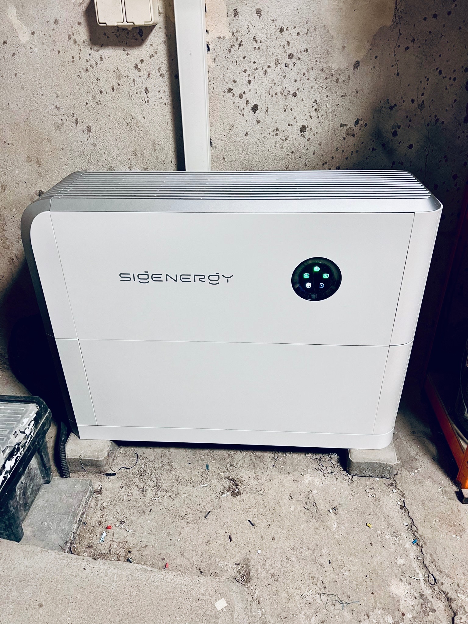 Biały akumulator energii SiGenergy z zielonymi diodami, umieszczony na betonowych bloczkach w surowym pomieszczeniu.