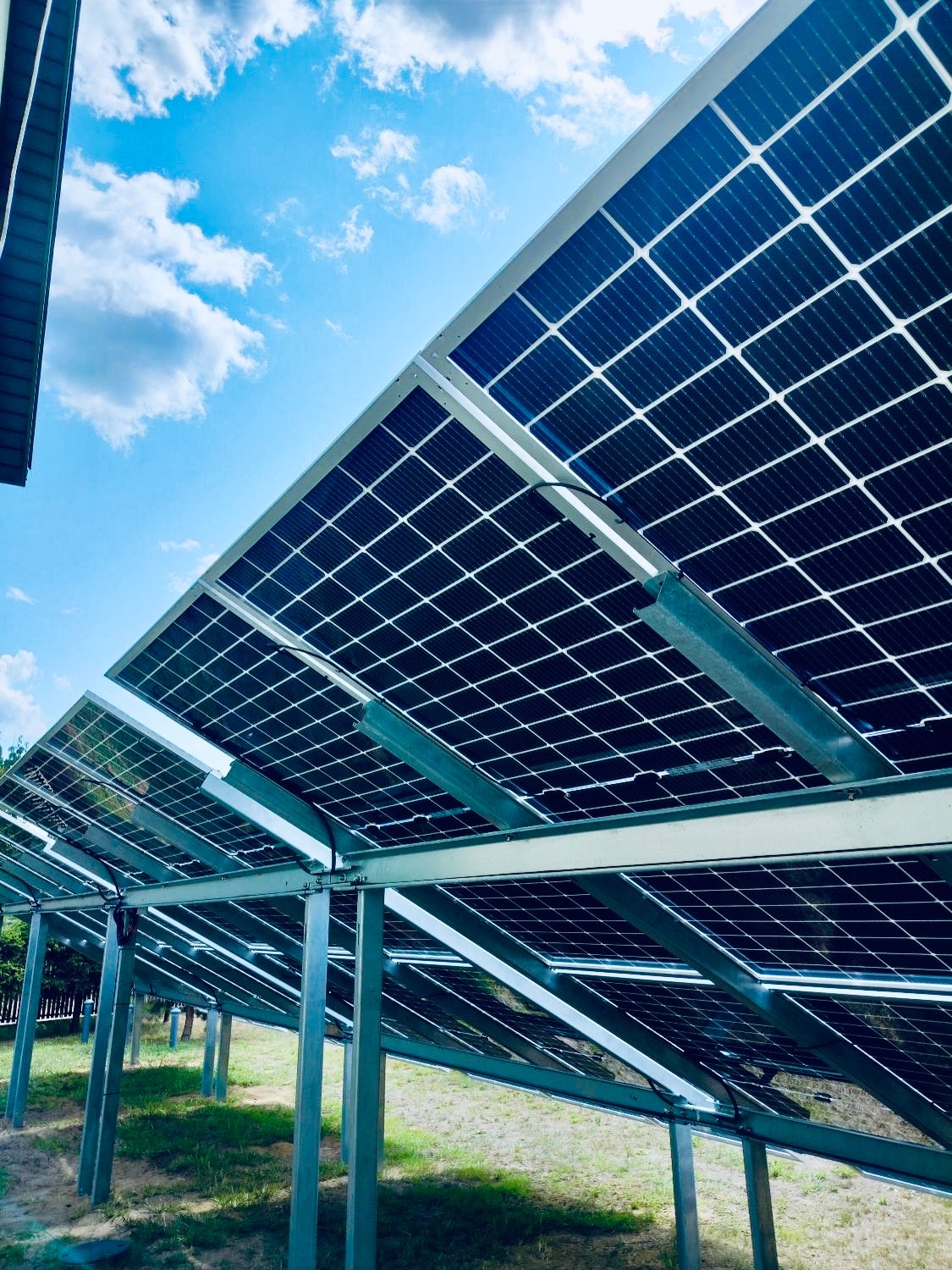 Instalacja paneli słonecznych z perspektywy gruntu, widoczne metalowe konstrukcje wsporcze na tle błękitnego nieba z chmurami, częściowo zacienione panele.