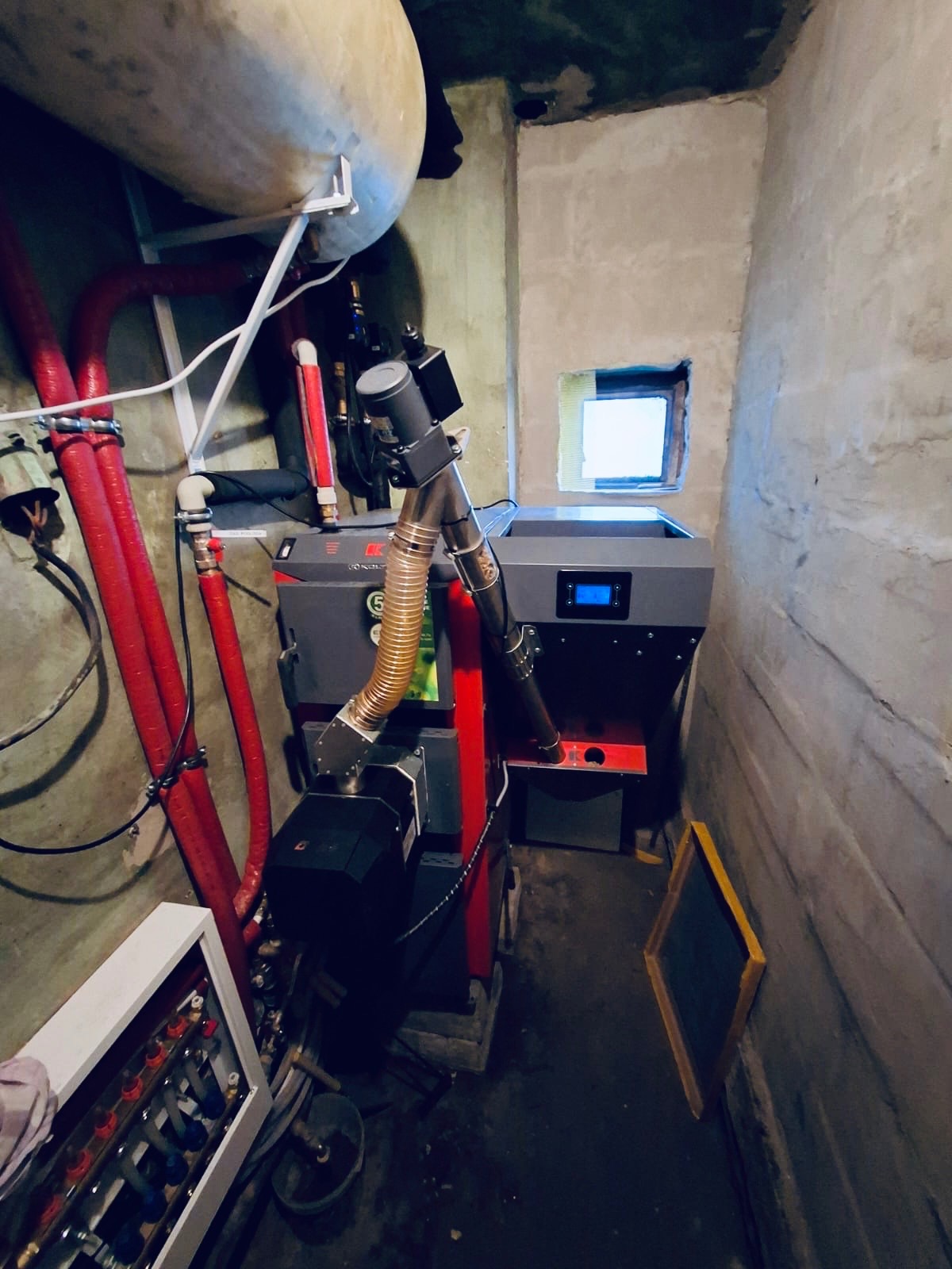 Nowoczesny piec na pellet w kotłowni z rurami grzewczymi i zasobnikiem, widoczny przez małe okienko. Ściany surowe, betonowe.