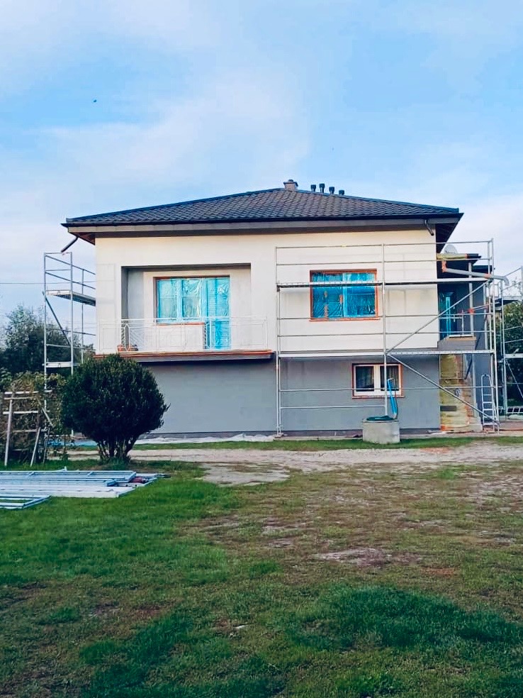 Dwukondygnacyjny dom w trakcie prac elewacyjnych, otoczony rusztowaniami, z oknami zabezpieczonymi folią, na tle zielonego trawnika i błękitnego nieba.