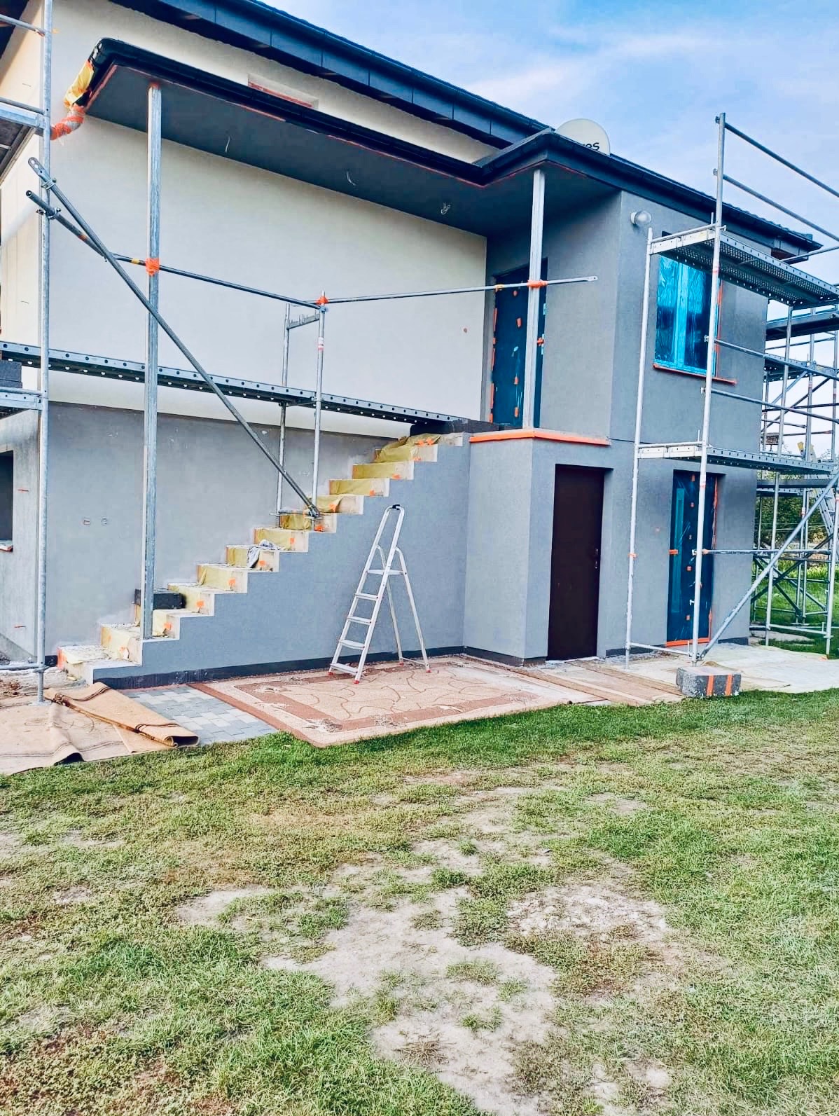 Dom w trakcie ocieplania: szare ściany, rusztowania, ocieplone schody wejściowe. Widoczne zabezpieczone okna i drzwi. Prace w toku.