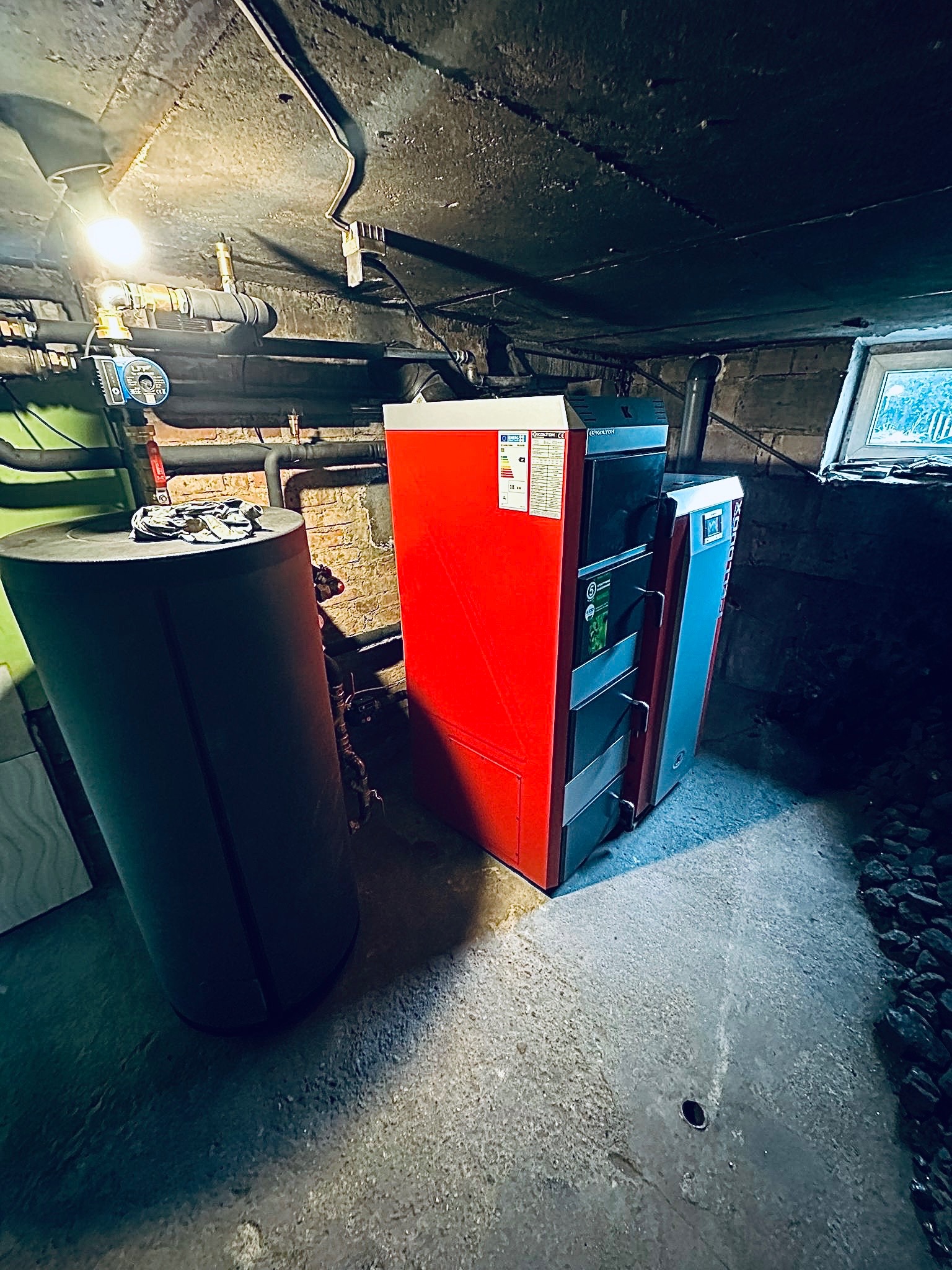 Czerwony piec na paliwo stałe marki Kolton obok zbiornika akumulacyjnego w kotłowni. Widoczne rury, instalacja, okno i węgiel.