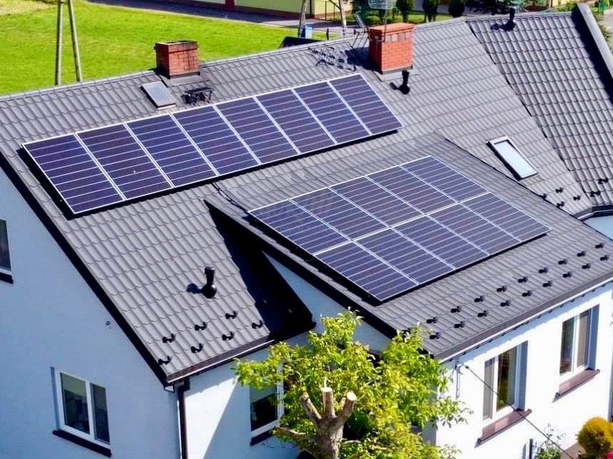 Panele fotowoltaiczne na szarym dachu dwuspadowym białego domu z kominami. Widok z góry, zielone otoczenie.