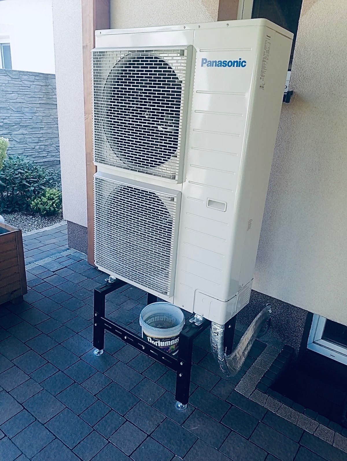 Biała pompa ciepła Panasonic na czarnym stojaku, obok ściany budynku z oknem. Widoczny fragment kostki brukowej i roślinności w tle.