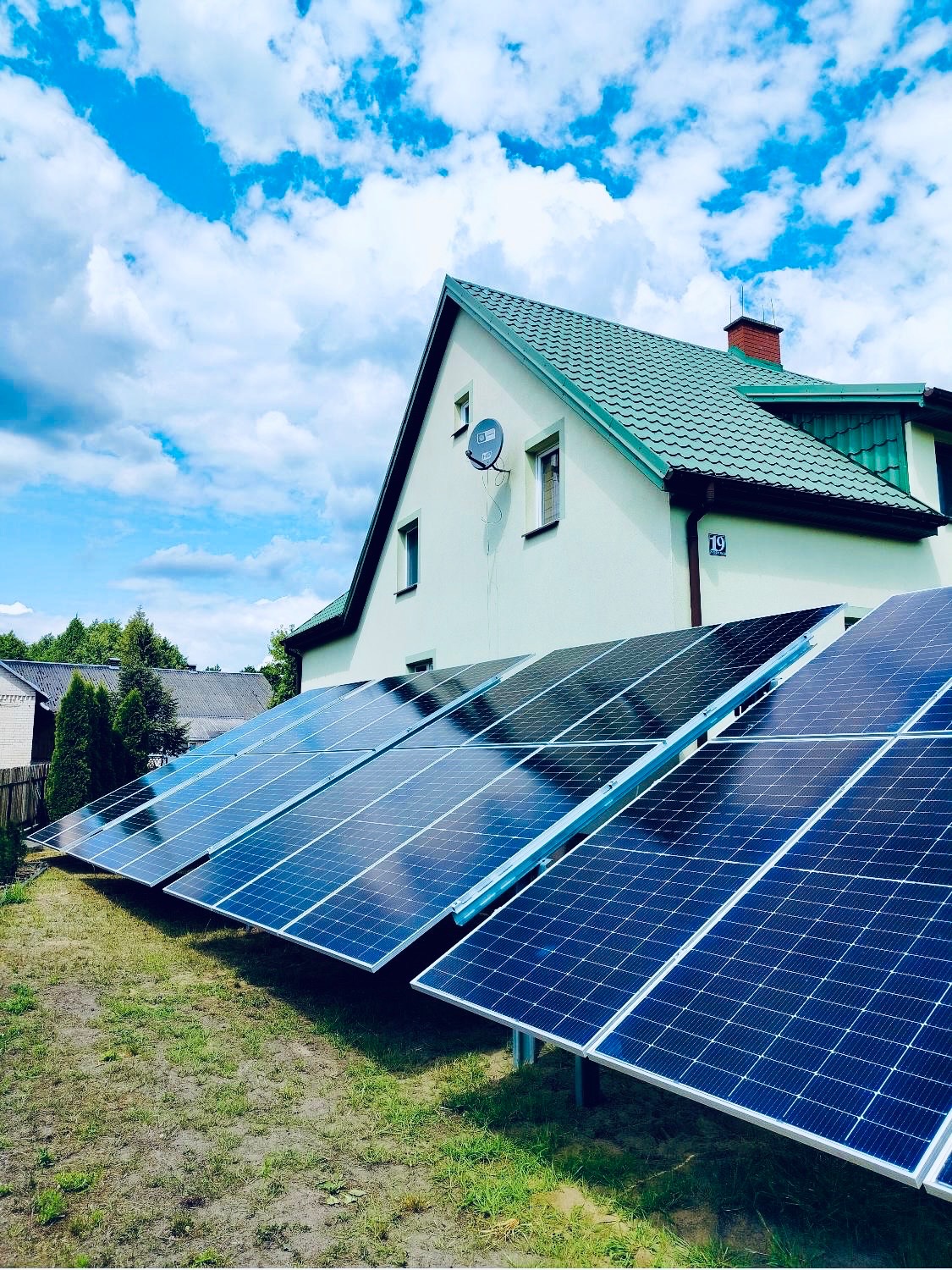 Instalacja paneli słonecznych na trawniku przed domem z zielonym dachem i anteną satelitarną. Błękitne niebo z chmurami w tle. Numer domu '19' widoczny na elewacji.