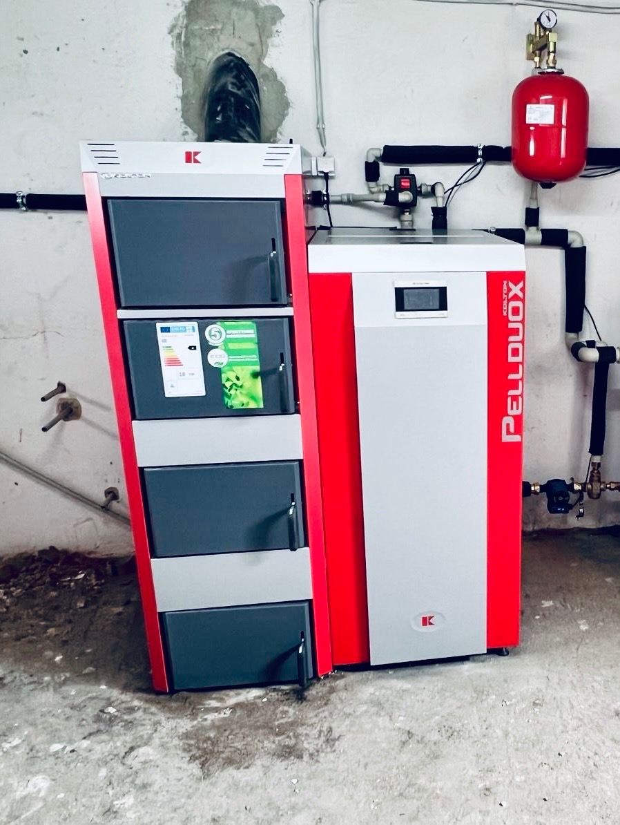 Kocioł na pellet Kolton PellDuoX z zasobnikiem, instalacja grzewcza z rurami w izolacji, zbiornik wyrównawczy. Montaż w kotłowni, widoczne przyłącza kominowe.