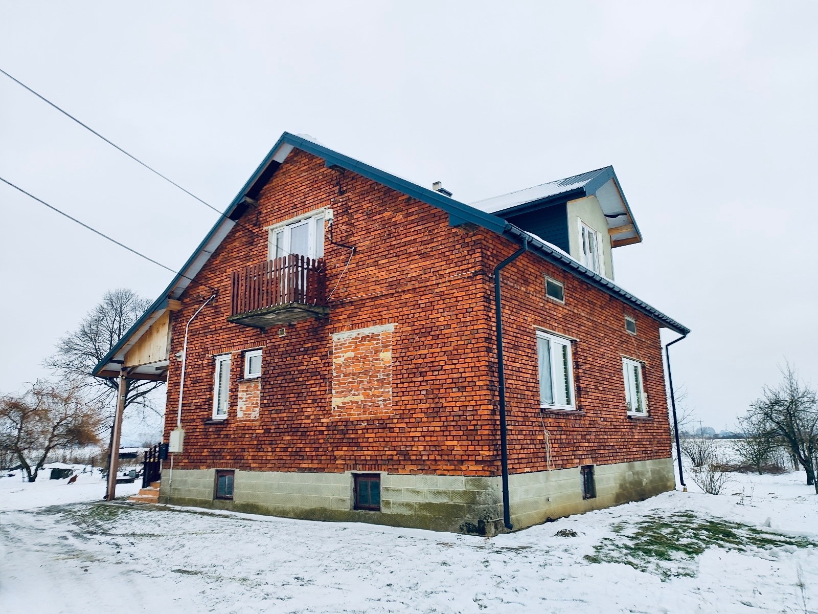 Ceglany dom z balkonem w zimowej scenerii. Widoczne ślady prac murarskich na ścianie. Białe okna kontrastują z czerwoną cegłą. Śnieg na trawniku i drzewach.