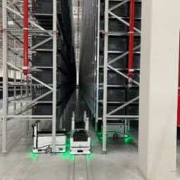 Automatyczny system magazynowy: regały z pojemnikami, roboty transportowe z zielonym oświetleniem w hali. Widok w głąb korytarza.