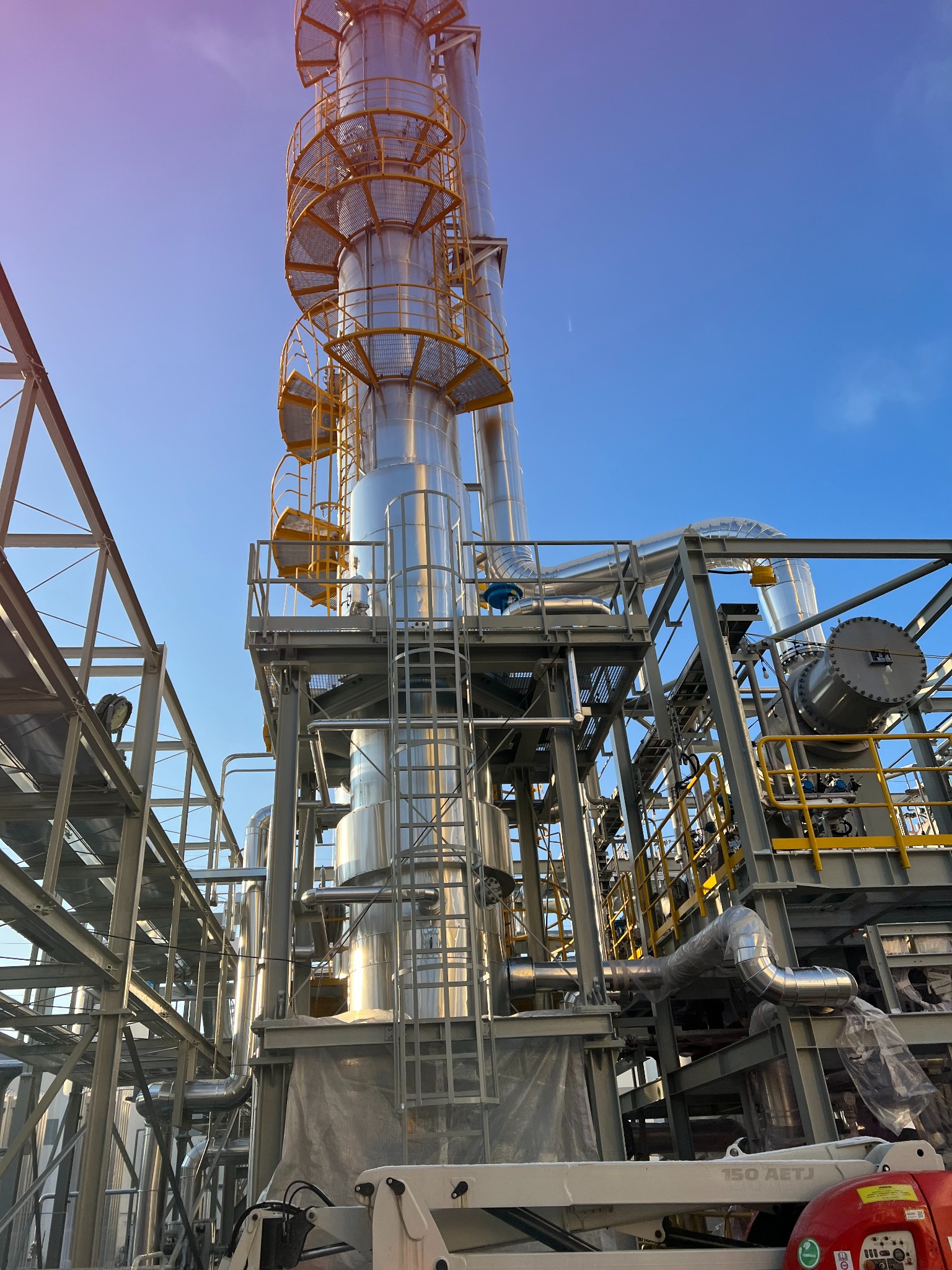 Wysoka, metalowa konstrukcja przemysłowa z rurami, schodami i platformami, wznosząca się na tle błękitnego nieba, widoczna z dołu. Elementy rusztowania i drabiny.