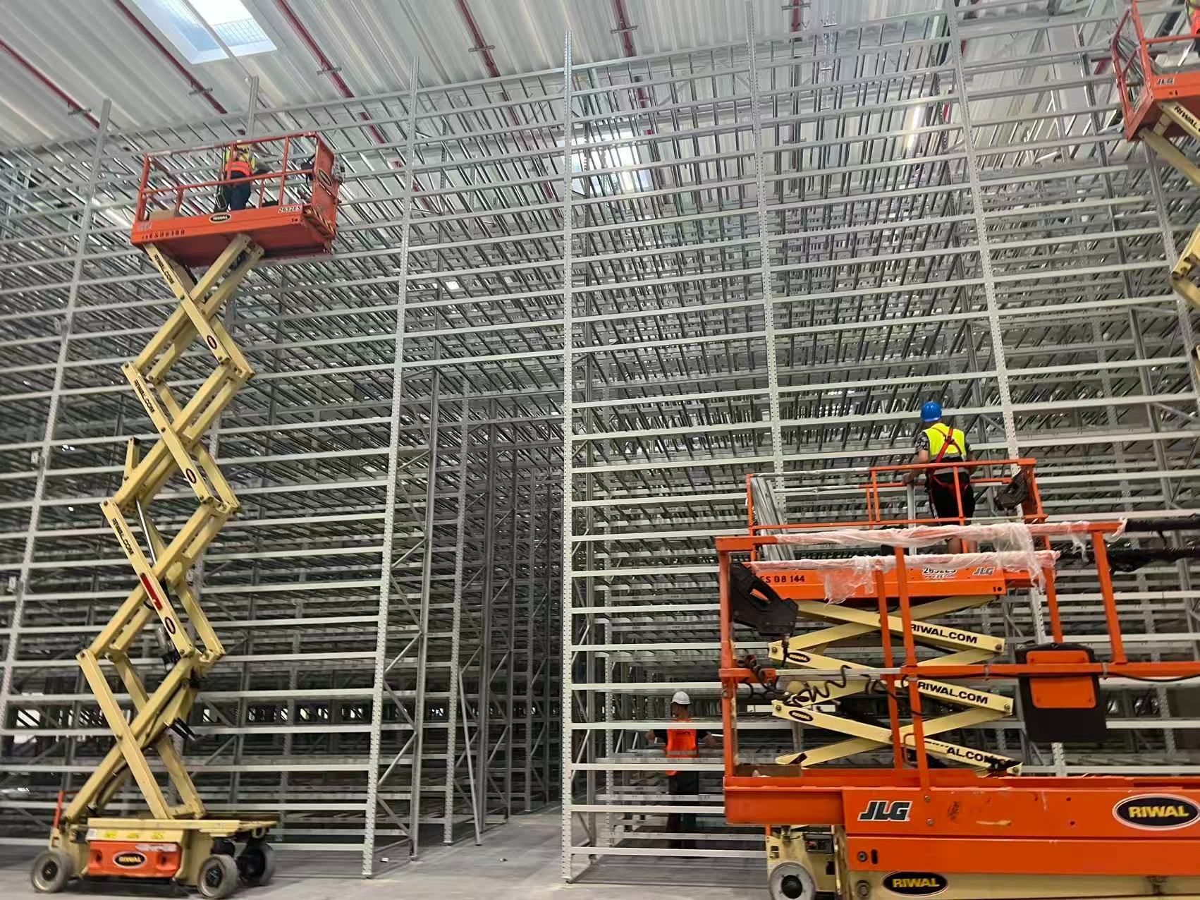 Montaż regałów magazynowych w hali: pracownicy na podnośnikach montują stalową konstrukcję regałów, widoczne elementy mocujące i oświetlenie.