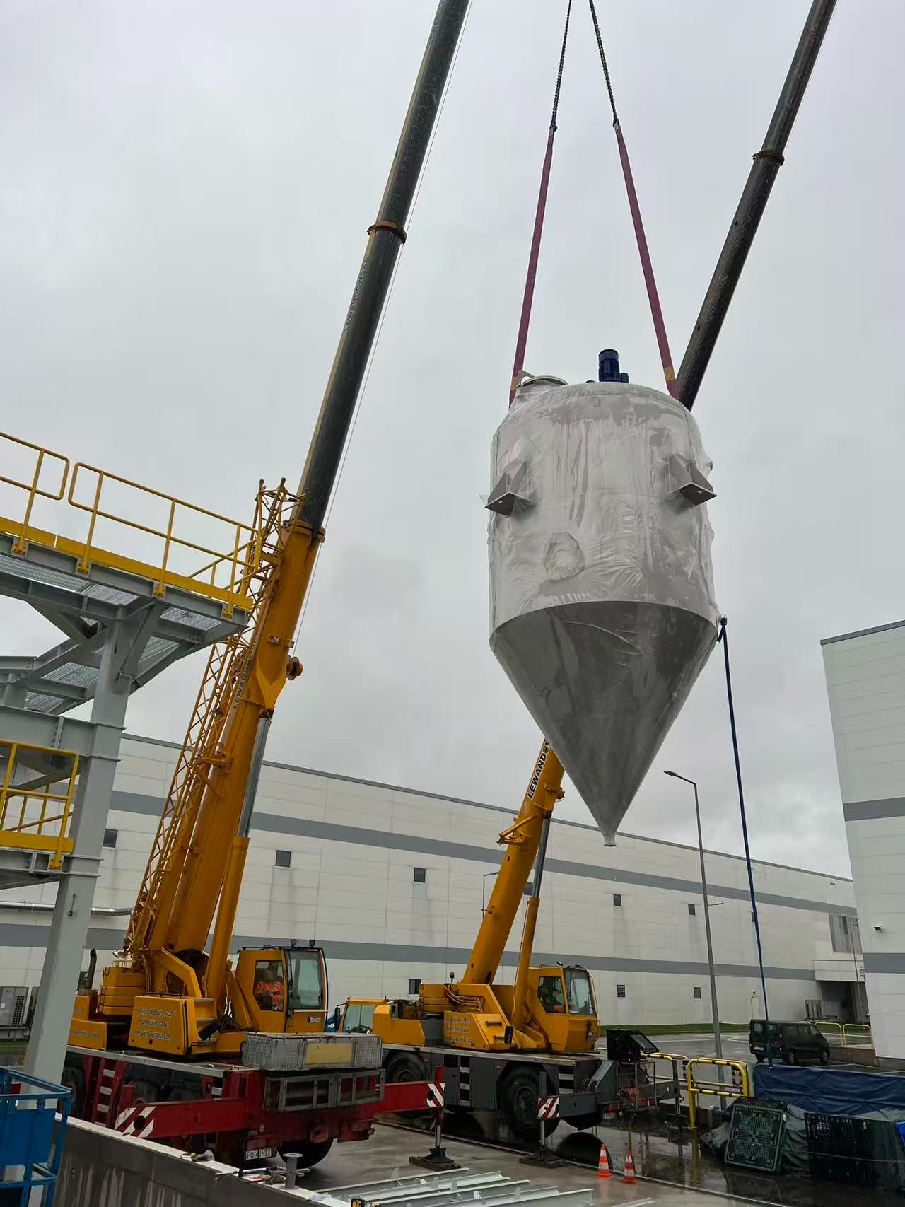 Montaż silosu na placu budowy hali magazynowej przy użyciu dwóch dźwigów, w tle budynek hali z szarej blachy. Silos owinięty folią ochronną.