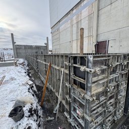 Sinaj Serhii Lylyk - Szare szalunki fundamentowe z metalowymi zaciskami, częściowo zasypane śniegiem, na tle betonowego budynku w pochmurny dzień. Widoczne zbrojenia i narzędzia.