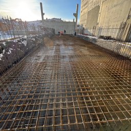 Sinaj Serhii Lylyk - Zbrojenie fundamentu z widoczną siatką stalową, przygotowane do zalania betonem. W tle fragment budynku w surowym stanie i pracownicy budowlani.