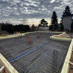 Sinaj Serhii Lylyk - Zbrojenie fundamentów pod budynek, widoczne szalunki z izolacją, wystające pręty zbrojeniowe i instalacje. Pochmurne niebo w tle.