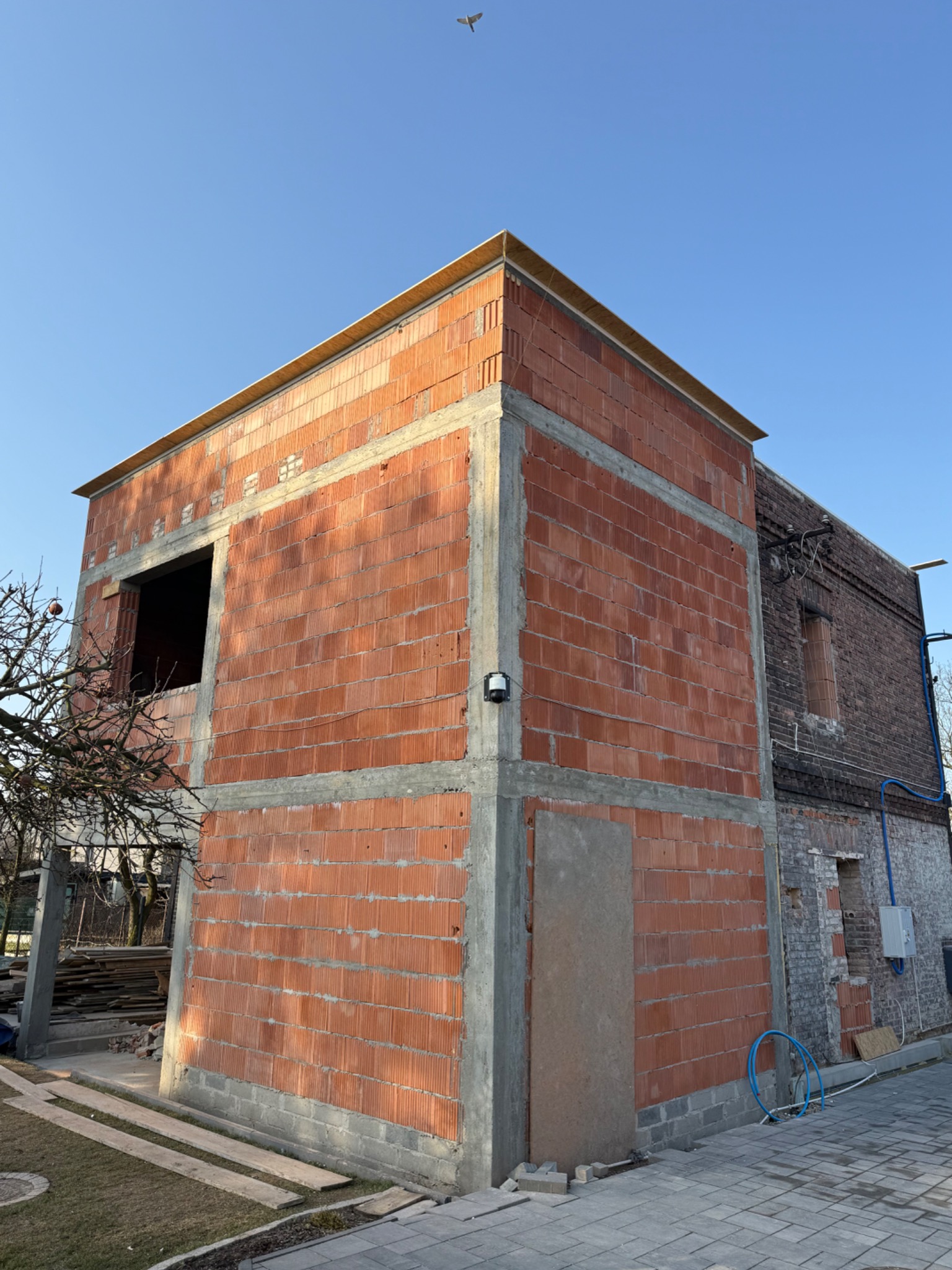Dwukondygnacyjny budynek w trakcie budowy z czerwonej cegły, z widocznymi betonowymi wzmocnieniami, na tle błękitnego nieba i starszego budynku z cegły.