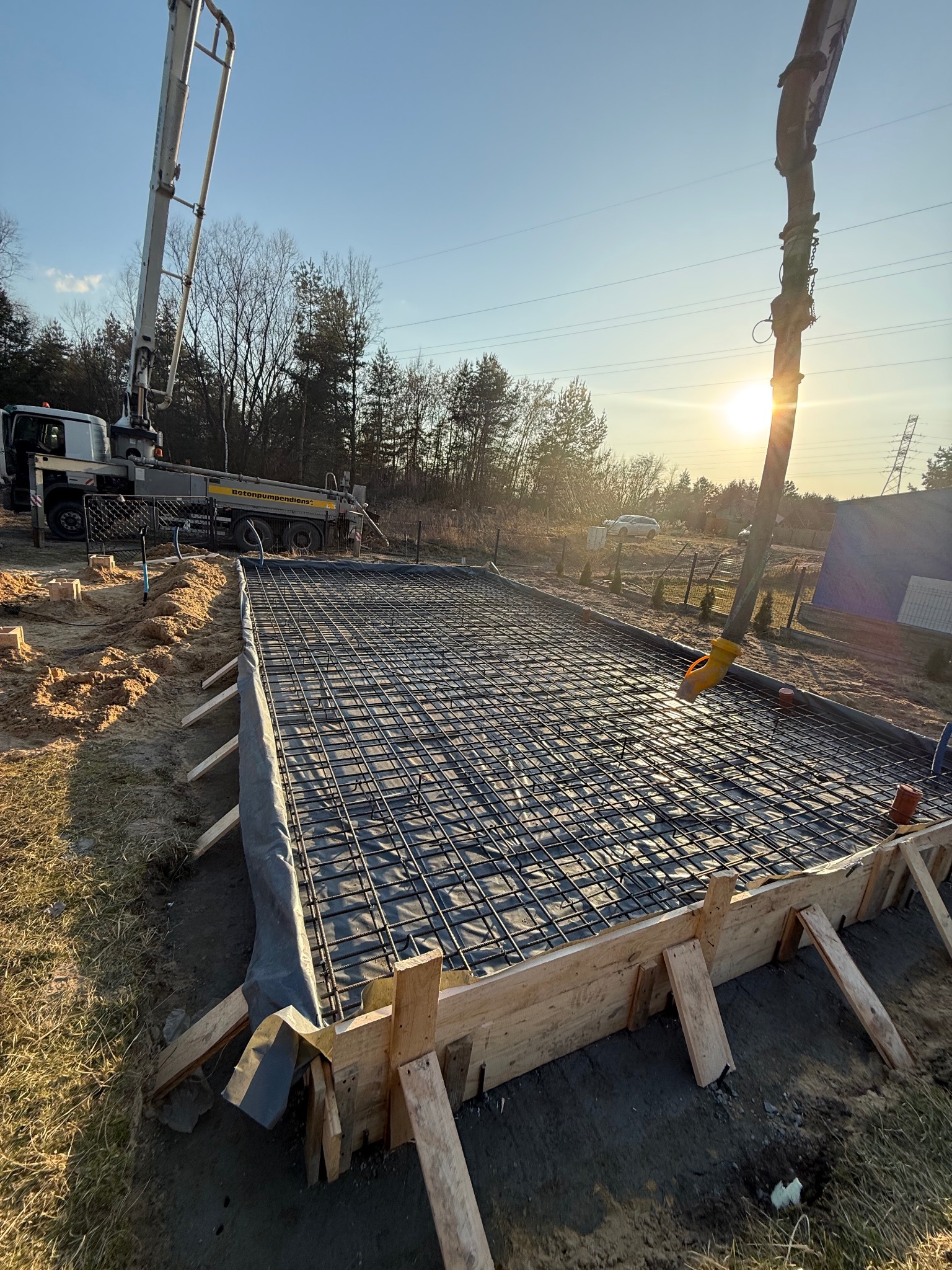 Zbrojenie fundamentów z szalunkiem i pompą do betonu w tle, oświetlone promieniami zachodzącego słońca. Widok na przygotowany teren pod wylewkę.