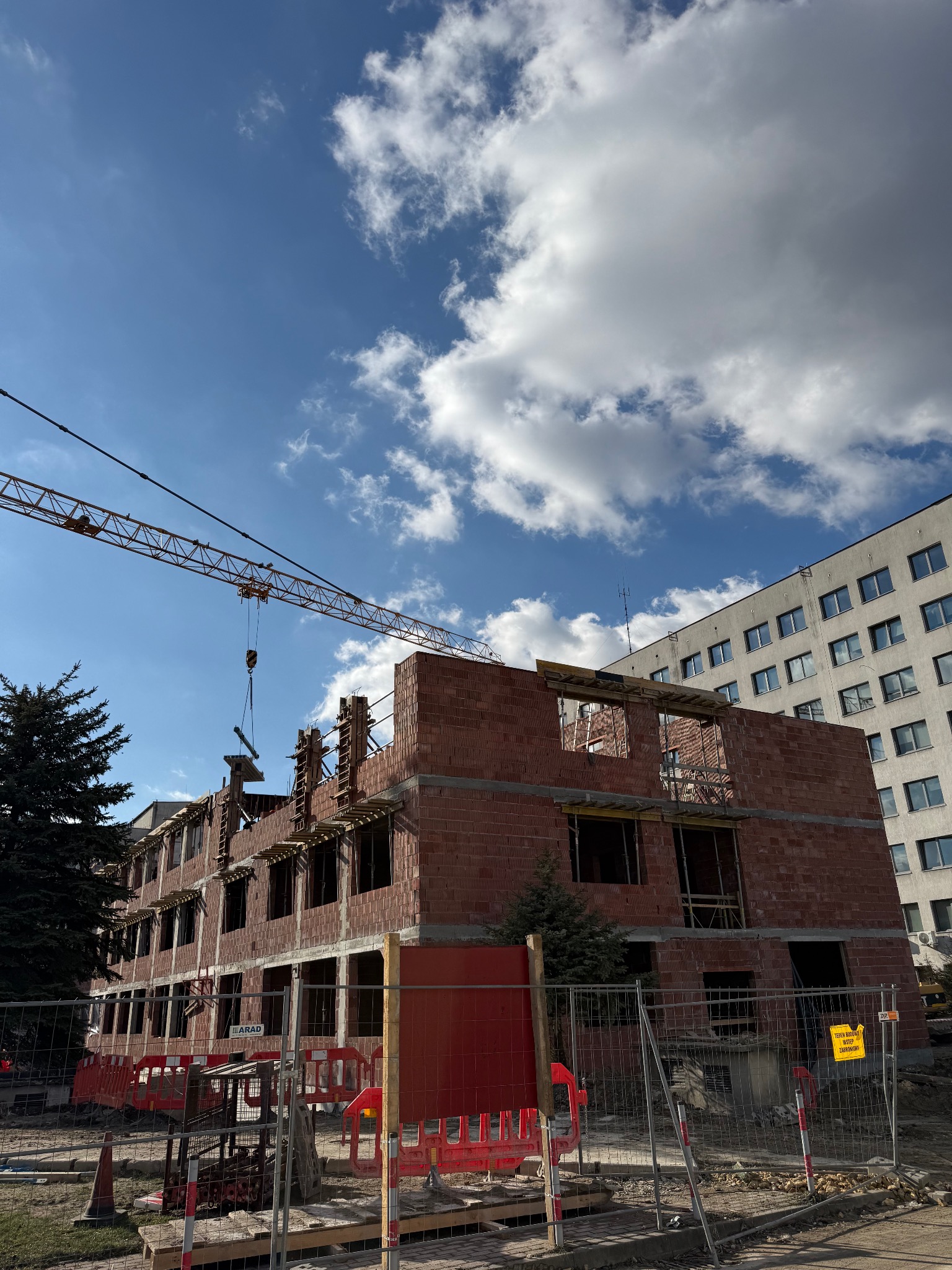 Wznoszenie dwupiętrowego budynku z czerwonej cegły w słoneczny dzień. Żuraw budowlany w tle, widoczne szalunki okienne. Częściowo ogrodzony teren budowy.