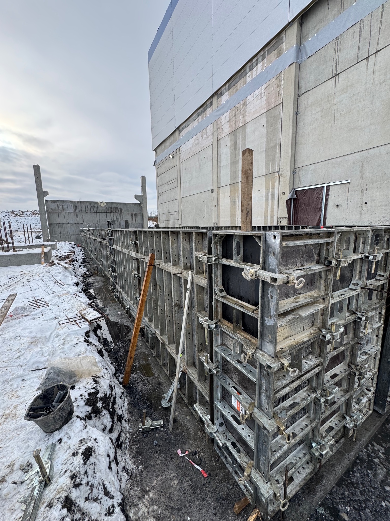 Szare szalunki fundamentowe z metalowymi zaciskami, częściowo zasypane śniegiem, na tle betonowego budynku w pochmurny dzień. Widoczne zbrojenia i narzędzia.