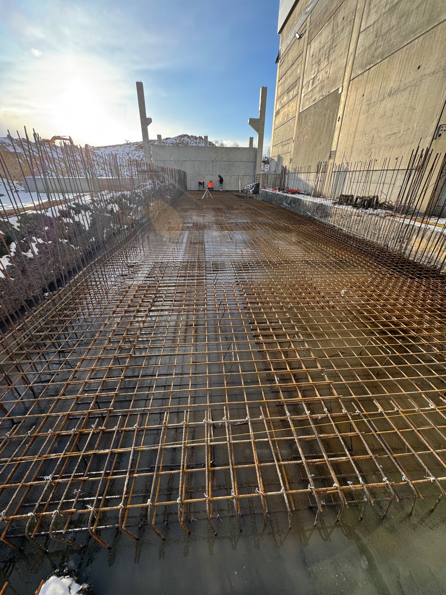 Zbrojenie fundamentu z widoczną siatką stalową, przygotowane do zalania betonem. W tle fragment budynku w surowym stanie i pracownicy budowlani.