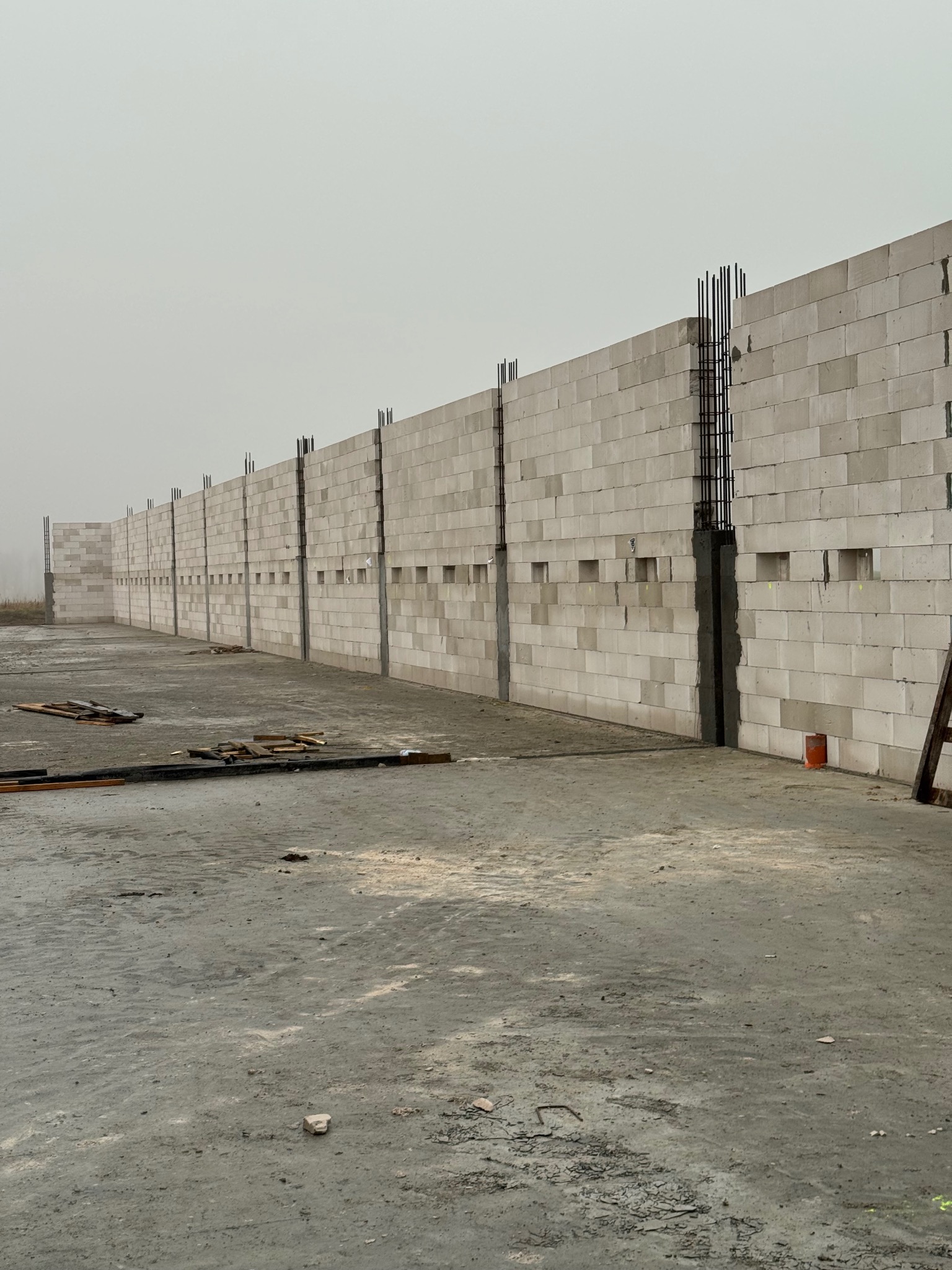 Wznoszenie ściany z betonu komórkowego z widocznym zbrojeniem pionowym. Szara, pochmurna pogoda. Na pierwszym planie betonowa posadzka i resztki materiałów budowlanych.