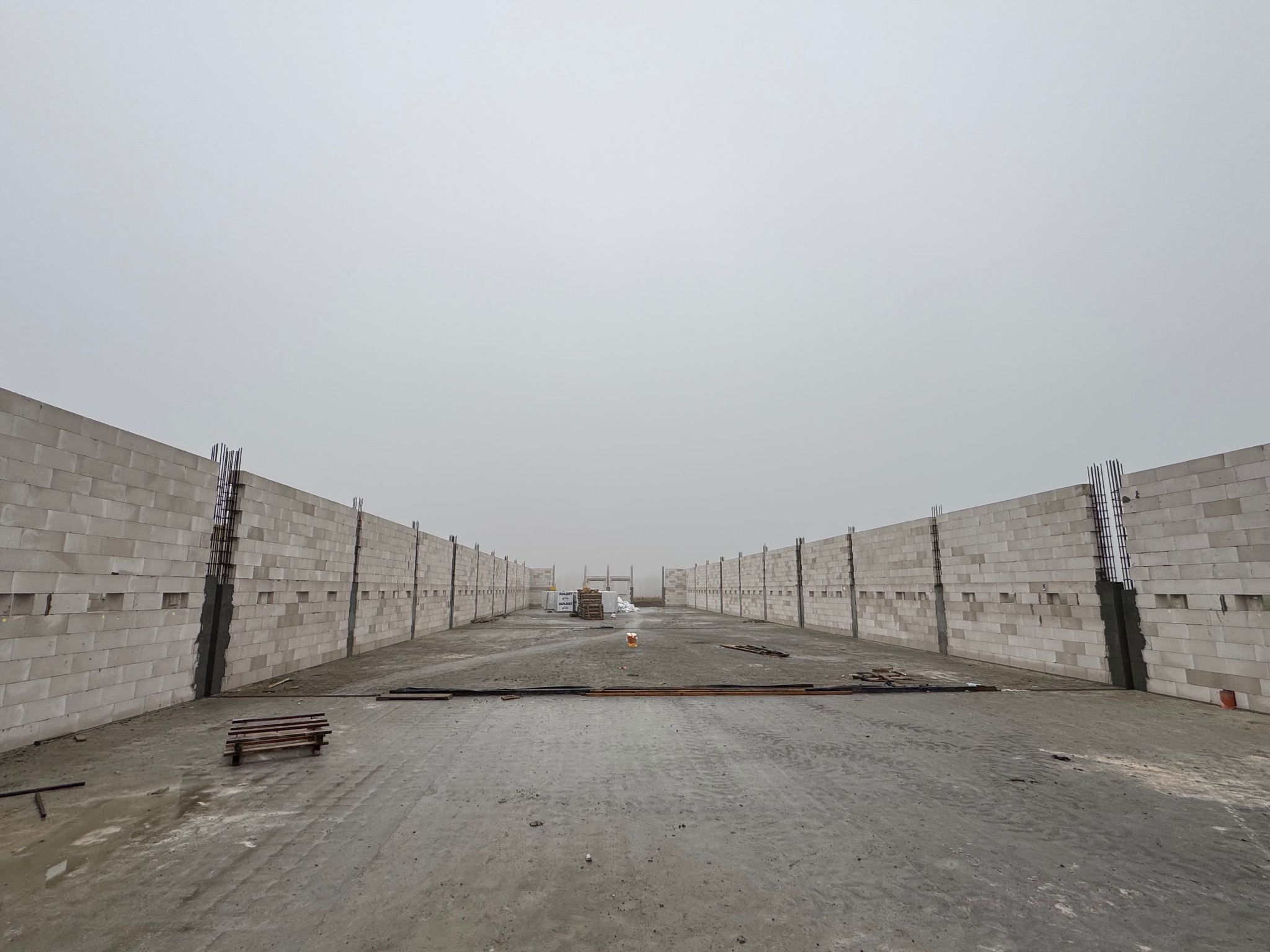 Wnętrze budowy hali: wzniesione ściany z betonu komórkowego z widocznym zbrojeniem, surowy beton na podłożu, mglista pogoda w tle. Etap murowania ścian.