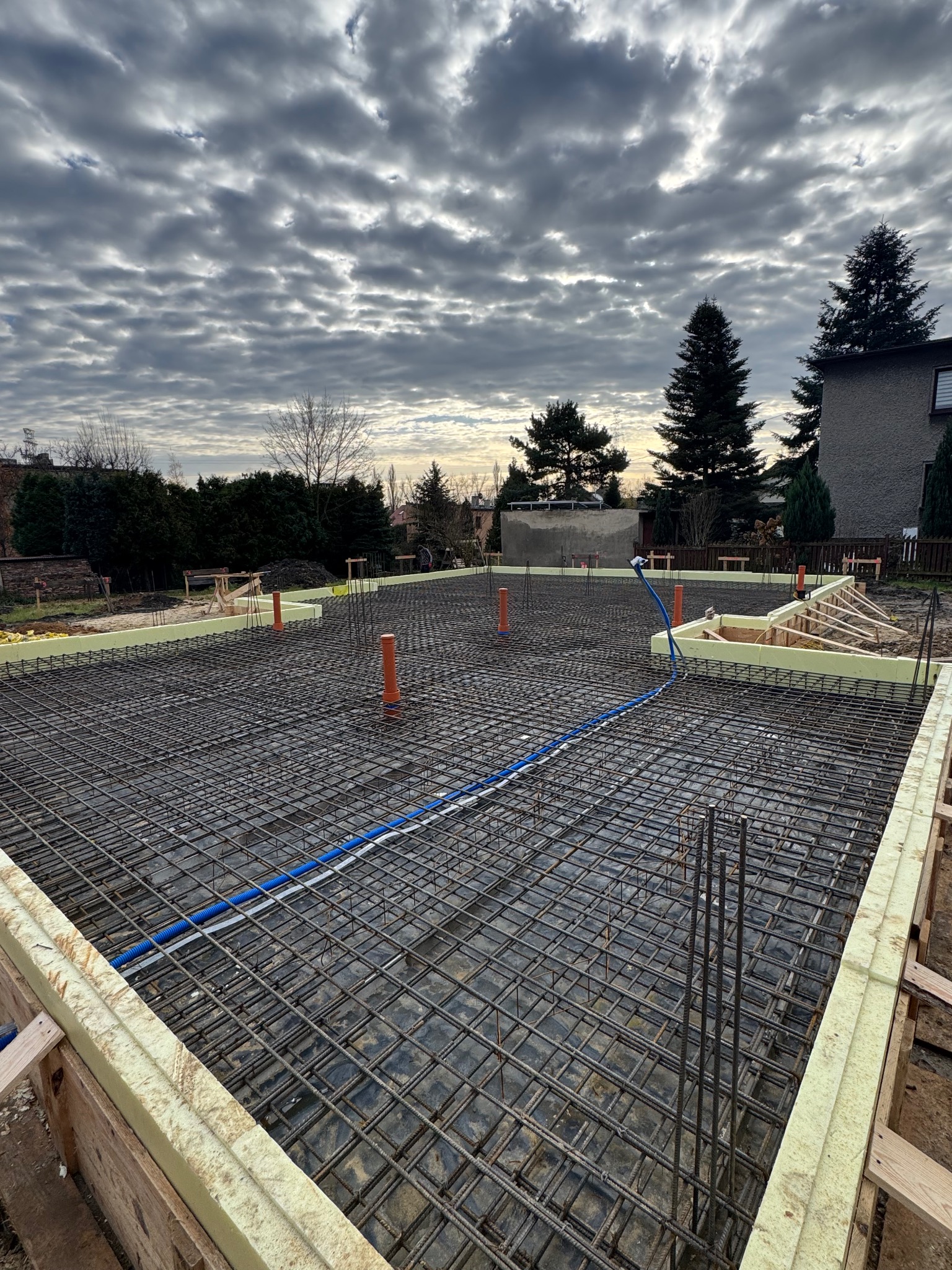 Zbrojenie fundamentów pod budynek, widoczne szalunki z izolacją, wystające pręty zbrojeniowe i instalacje. Pochmurne niebo w tle.