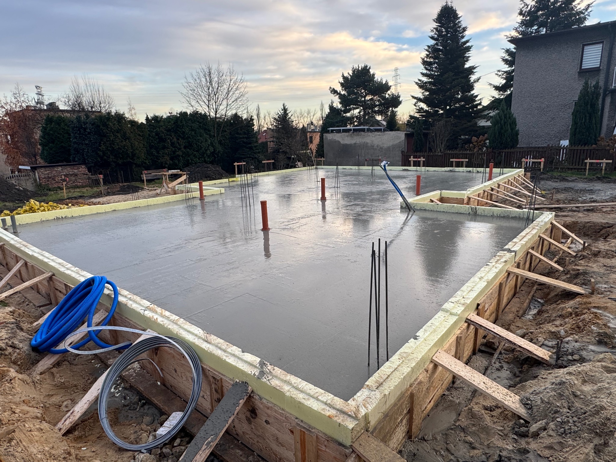 Zalany betonem fundament domu jednorodzinnego, z widocznym szalunkiem, izolacją termiczną i wystającymi prętami zbrojeniowymi. W tle zabudowa jednorodzinna i pochmurne niebo.