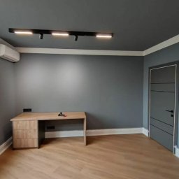 M&K - Wnętrze pokoju po malowaniu: szare ściany i sufit, jasna podłoga, drewniane biurko. Widoczna listwa oświetleniowa i klimatyzator. Nowoczesne wykończenie.