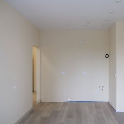 M&K - Jasne, świeżo pomalowane wnętrze z drewnianą podłogą i przygotowanymi instalacjami. Widoczne punkty świetlne i gniazdka elektryczne, gotowe do dalszego wykończenia.