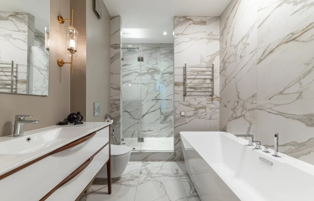 Elegancka łazienka z marmurowymi płytkami, kabiną prysznicową i wolnostojącą wanną. Umywalka z szafką i złote akcenty oświetleniowe dodają luksusowego charakteru.