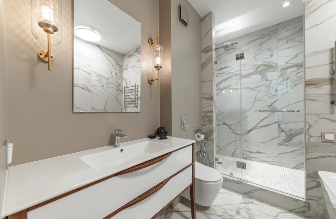 Elegancka łazienka z marmurowymi płytkami, kabiną prysznicową i białymi meblami z drewnianymi akcentami. Oświetlenie punktowe i złote lampy ścienne.