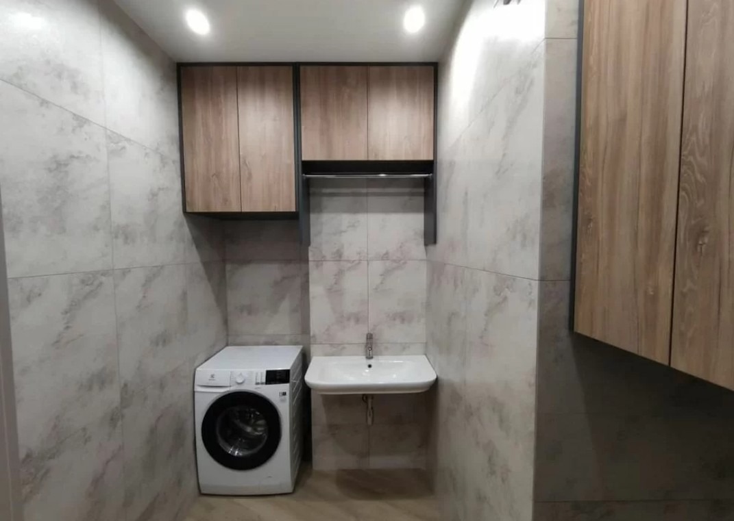 Wykończona łazienka z pralką, umywalką i szafkami w odcieniach drewna i szarości. Płytki imitują marmur. Minimalistyczny design.