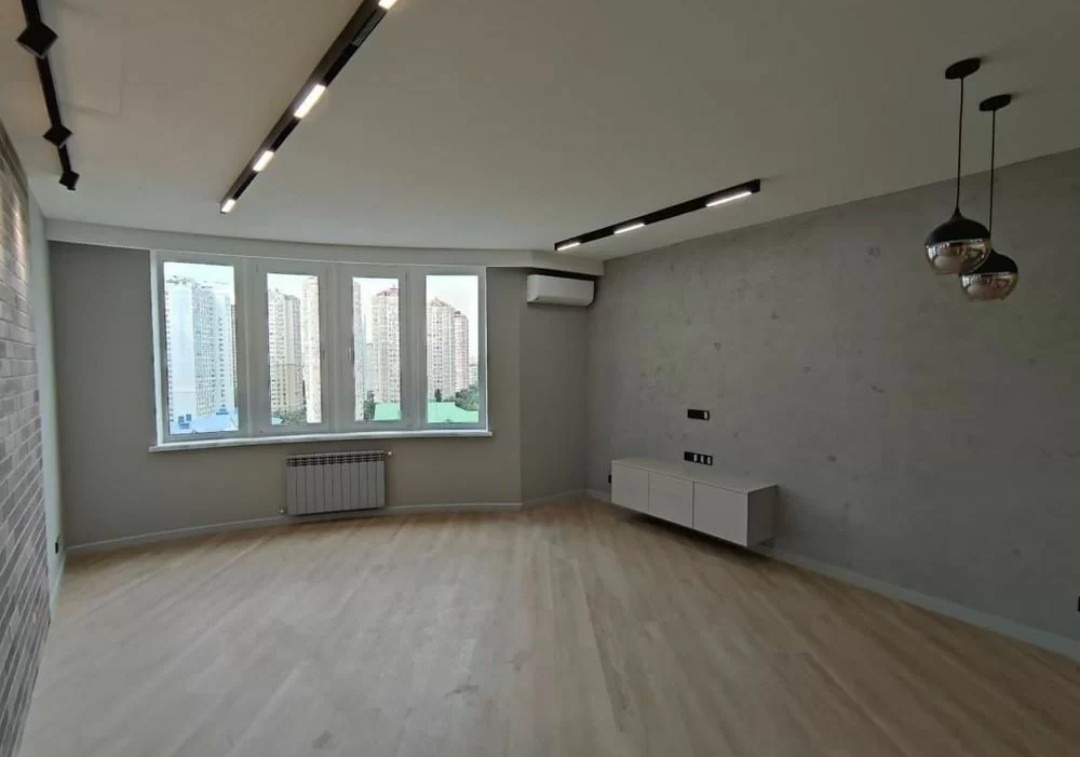 Jasne, przestronne wnętrze po remoncie z parkietem, szarą ścianą i oknem z widokiem na miasto. Nowoczesne oświetlenie sufitowe i wiszące lampy dodają elegancji.