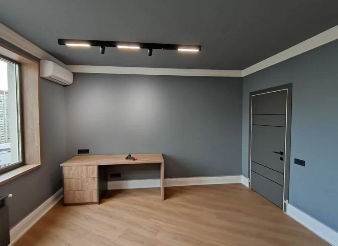 Wnętrze pokoju po malowaniu: szare ściany i sufit, jasna podłoga, drewniane biurko. Widoczna listwa oświetleniowa i klimatyzator. Nowoczesne wykończenie.